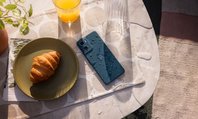 O smartphone Motorola Moto G06 na cor azul marinho, molhado, deitado em uma mesa. Ao lado do aparelho, há um croissant em um prato e um copo de suco.