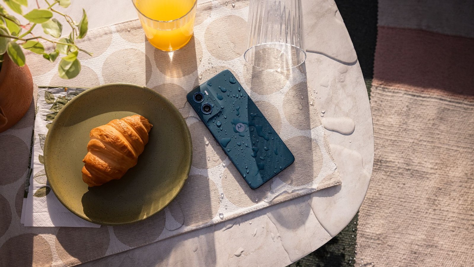 O smartphone Motorola Moto G06 na cor azul marinho, molhado, deitado em uma mesa. Ao lado do aparelho, há um croissant em um prato e um copo de suco.