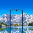 Um smartphone Samsung Galaxy A17 com um entalhe em forma de gota d'água está centralizado em primeiro plano. A tela do telefone exibe um prédio com fachada amarela e múltiplas janelas, cercado por árvores. O prédio parece estar localizado próximo a um corpo de água, e seu reflexo é visível na tela. O fundo da imagem é uma paisagem deslumbrante, com um grande e calmo lago em primeiro plano e uma cordilheira com picos nevados sob um céu azul brilhante ao fundo. A tela do celular aparece mostrando uma vista ampliada e recortada do prédio e de seus arredores imediatos a partir da paisagem do fundo, criando um efeito de moldura dentro de uma moldura. A imagem geral possui uma paleta de cores vibrante e aprimorada.