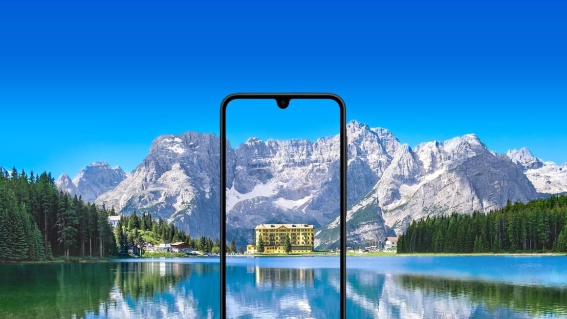 Um smartphone Samsung Galaxy A17 com um entalhe em forma de gota d'água está centralizado em primeiro plano. A tela do telefone exibe um prédio com fachada amarela e múltiplas janelas, cercado por árvores. O prédio parece estar localizado próximo a um corpo de água, e seu reflexo é visível na tela. O fundo da imagem é uma paisagem deslumbrante, com um grande e calmo lago em primeiro plano e uma cordilheira com picos nevados sob um céu azul brilhante ao fundo. A tela do celular aparece mostrando uma vista ampliada e recortada do prédio e de seus arredores imediatos a partir da paisagem do fundo, criando um efeito de moldura dentro de uma moldura. A imagem geral possui uma paleta de cores vibrante e aprimorada.