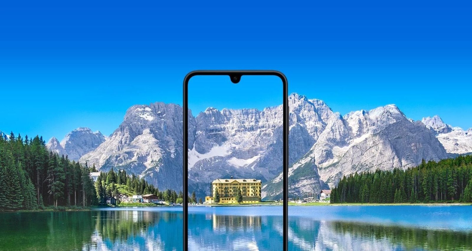 Um smartphone Samsung Galaxy A17 com um entalhe em forma de gota d'água está centralizado em primeiro plano. A tela do telefone exibe um prédio com fachada amarela e múltiplas janelas, cercado por árvores. O prédio parece estar localizado próximo a um corpo de água, e seu reflexo é visível na tela. O fundo da imagem é uma paisagem deslumbrante, com um grande e calmo lago em primeiro plano e uma cordilheira com picos nevados sob um céu azul brilhante ao fundo. A tela do celular aparece mostrando uma vista ampliada e recortada do prédio e de seus arredores imediatos a partir da paisagem do fundo, criando um efeito de moldura dentro de uma moldura. A imagem geral possui uma paleta de cores vibrante e aprimorada.