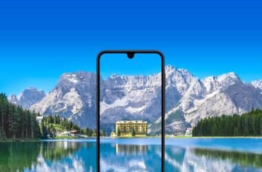 Um smartphone Samsung Galaxy A17 com um entalhe em forma de gota d'água está centralizado em primeiro plano. A tela do telefone exibe um prédio com fachada amarela e múltiplas janelas, cercado por árvores. O prédio parece estar localizado próximo a um corpo de água, e seu reflexo é visível na tela. O fundo da imagem é uma paisagem deslumbrante, com um grande e calmo lago em primeiro plano e uma cordilheira com picos nevados sob um céu azul brilhante ao fundo. A tela do celular aparece mostrando uma vista ampliada e recortada do prédio e de seus arredores imediatos a partir da paisagem do fundo, criando um efeito de moldura dentro de uma moldura. A imagem geral possui uma paleta de cores vibrante e aprimorada.