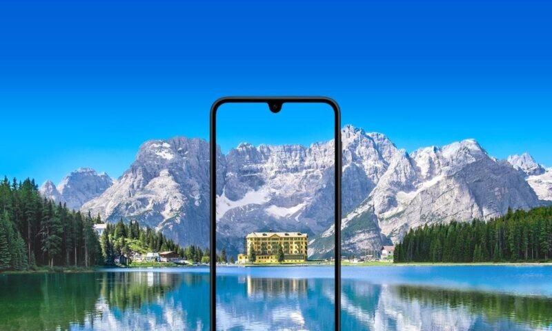 Um smartphone Samsung Galaxy A17 com um entalhe em forma de gota d'água está centralizado em primeiro plano. A tela do telefone exibe um prédio com fachada amarela e múltiplas janelas, cercado por árvores. O prédio parece estar localizado próximo a um corpo de água, e seu reflexo é visível na tela. O fundo da imagem é uma paisagem deslumbrante, com um grande e calmo lago em primeiro plano e uma cordilheira com picos nevados sob um céu azul brilhante ao fundo. A tela do celular aparece mostrando uma vista ampliada e recortada do prédio e de seus arredores imediatos a partir da paisagem do fundo, criando um efeito de moldura dentro de uma moldura. A imagem geral possui uma paleta de cores vibrante e aprimorada.