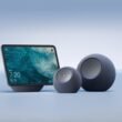 Três novos dispositivos da linha Amazon Echo 2025, todos na cor grafite e com acabamento em tecido. A imagem exibe o Echo Show com tela, o Echo Dot Max (esférico e menor) e o Echo Studio (esférico e maior).