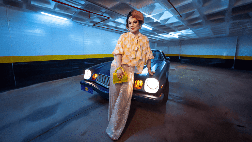 Lorelay Fox, drag queen com roupa de paetês, posando ao lado de um Chevrolet Camaro Z/28 azul de 1979 em uma garagem subterrânea com paredes azuis, amarelas e pretas