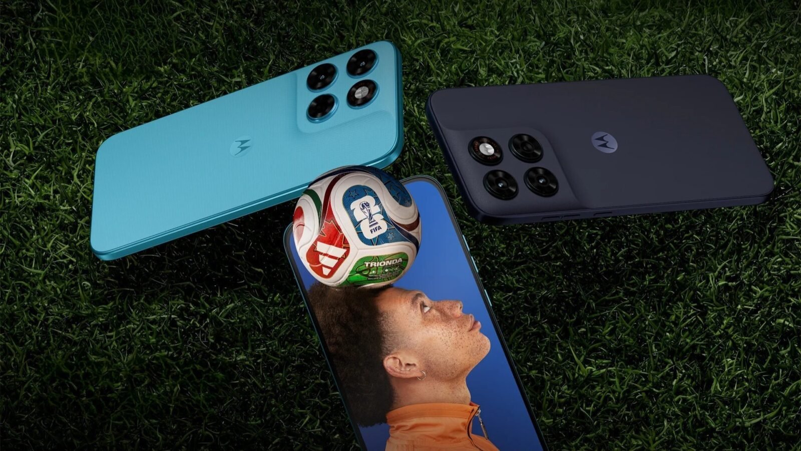 Três smartphones Motorola Moto G Max estão dispostos sobre um gramado verde escuro. Dois aparelhos aparecem de costas, destacando o módulo de câmeras quádruplo e o logotipo da marca no centro: um na cor azul claro, posicionado na diagonal superior esquerda, e outro na cor grafite, na diagonal superior direita. Centralizado na parte inferior, o terceiro aparelho está de frente, exibindo em sua tela a imagem de um jovem com uma bola de futebol equilibrada na cabeça sob um fundo azul.
