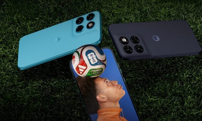 Três smartphones Motorola Moto G Max estão dispostos sobre um gramado verde escuro. Dois aparelhos aparecem de costas, destacando o módulo de câmeras quádruplo e o logotipo da marca no centro: um na cor azul claro, posicionado na diagonal superior esquerda, e outro na cor grafite, na diagonal superior direita. Centralizado na parte inferior, o terceiro aparelho está de frente, exibindo em sua tela a imagem de um jovem com uma bola de futebol equilibrada na cabeça sob um fundo azul.