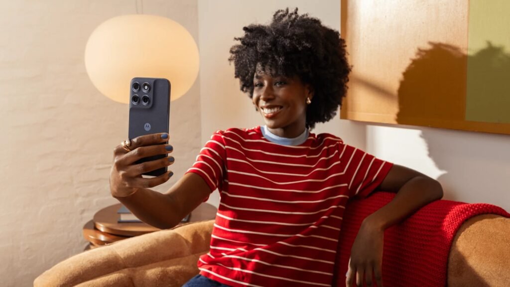 Uma mulher negra sorridente, com cabelo estilo black power, está sentada em um sofá e segura o smartphone Motorola Moto G Max na cor grafite para tirar uma selfie. Ela veste uma camiseta vermelha com listras horizontais brancas e gola redonda. O aparelho está posicionado verticalmente, mostrando sua traseira com o logotipo da Motorola e o módulo quadrado de câmeras quádruplas direcionado para ela. O ambiente interno tem iluminação calorosa e aconchegante, com uma luminária de mesa esférica acesa ao fundo à esquerda e uma manta vermelha sobre o encosto do sofá.